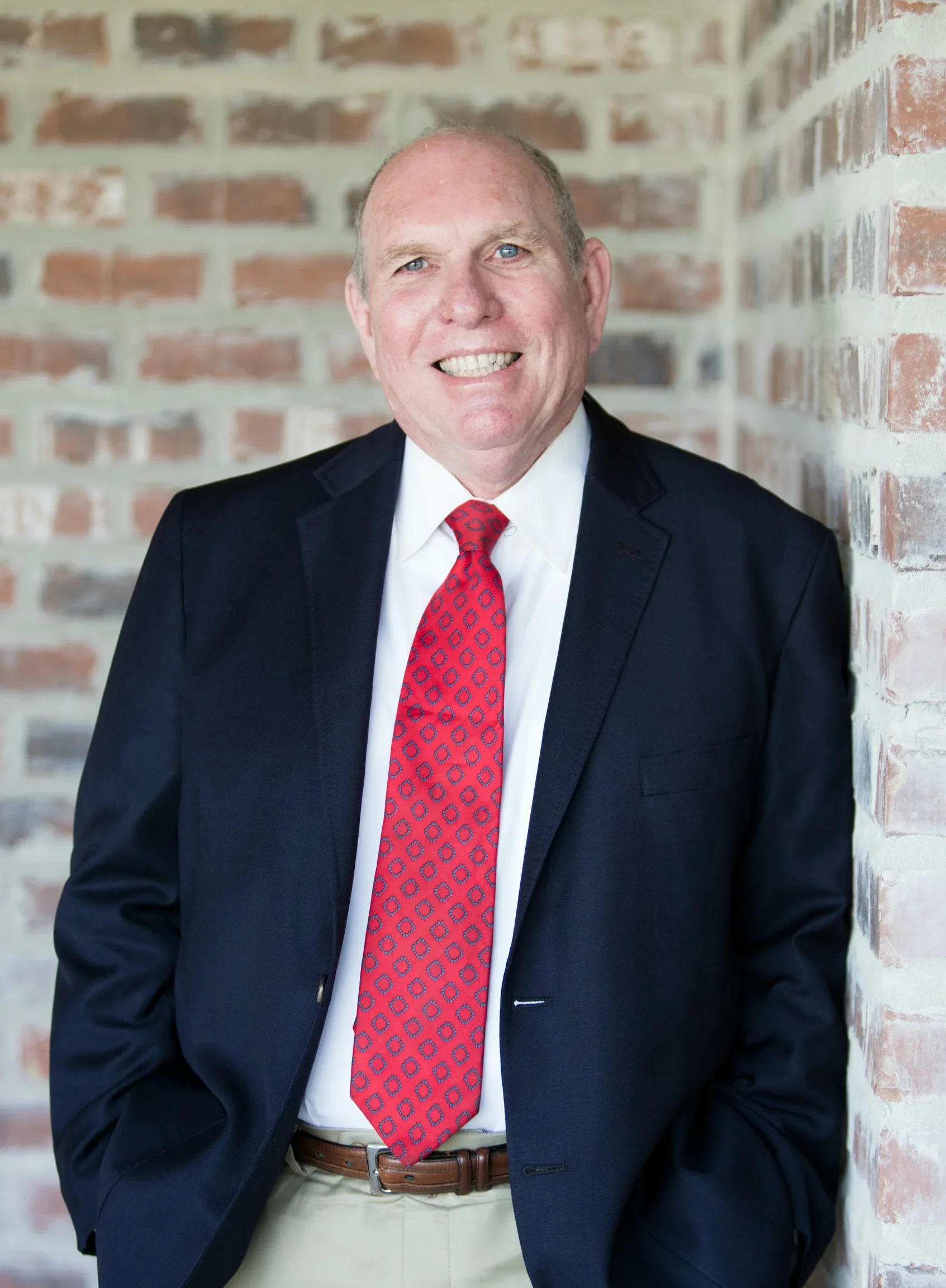 Smiling man in business suit with red patterned tie, standing against a brick wall.
