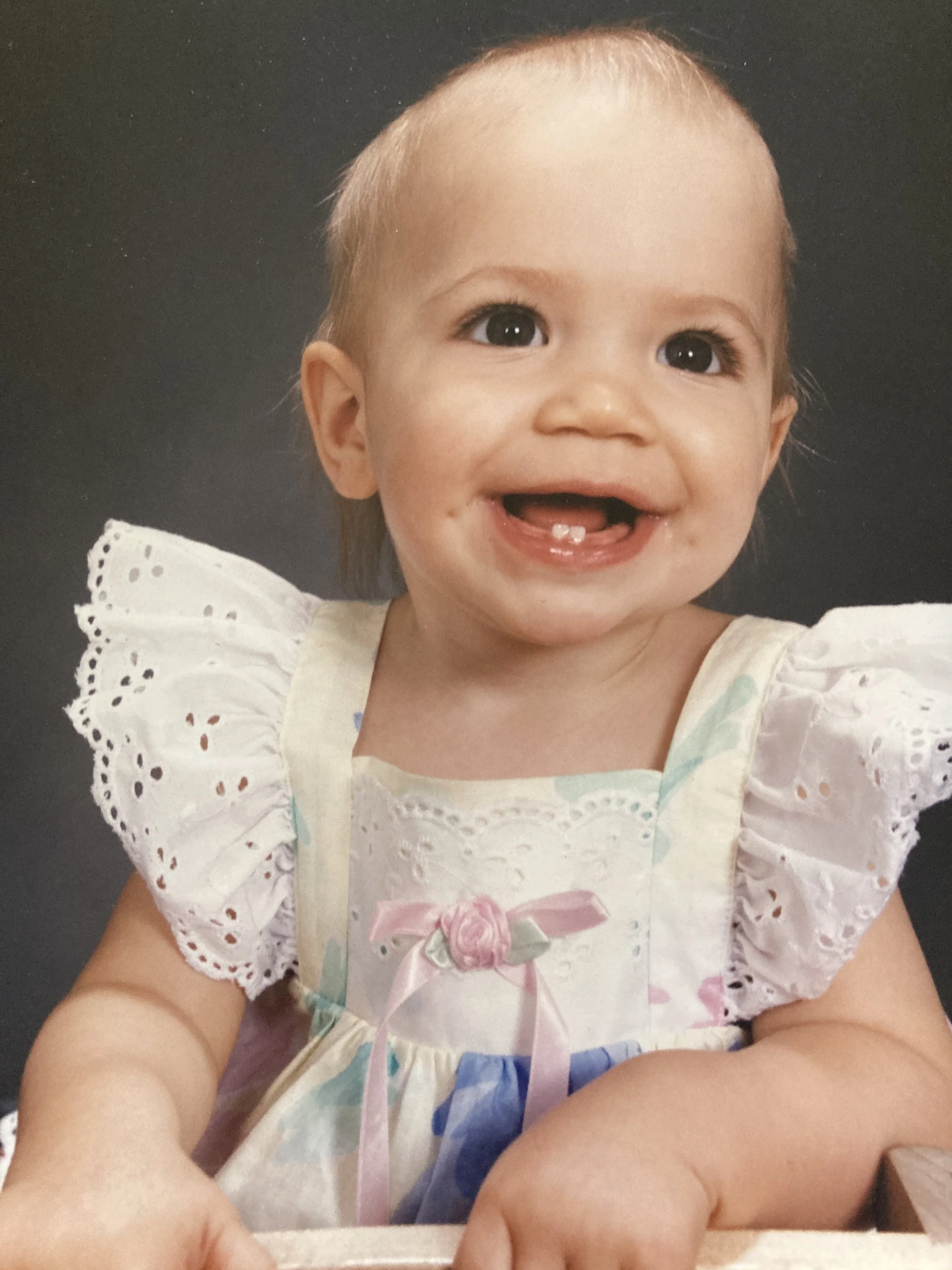 Dana as a smiling baby girl with light blonde hair and big dark eyes, wearing a pastel-colored dress with lace and ribbon details, sitting against a dark background.