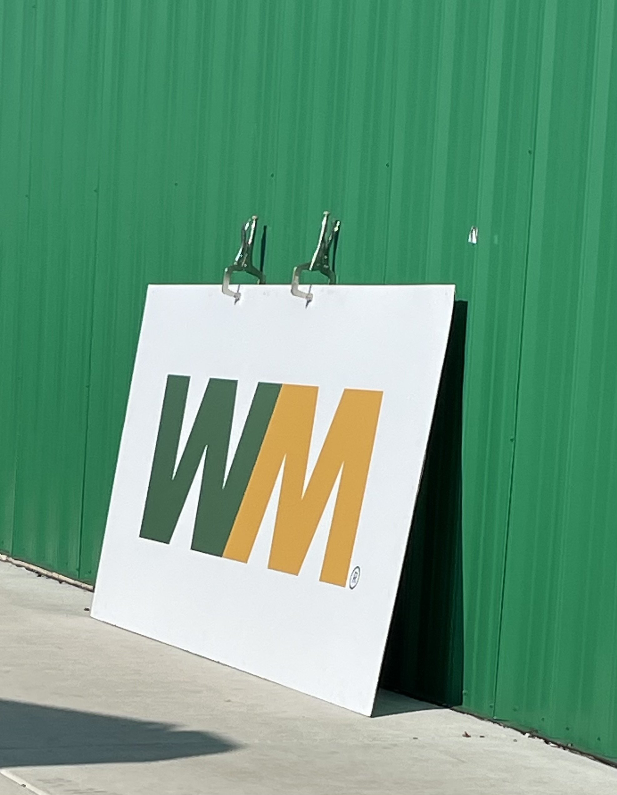 White sign with a logo reading 'WAM' in green, yellow, and black letters, leaning against a green metal wall.