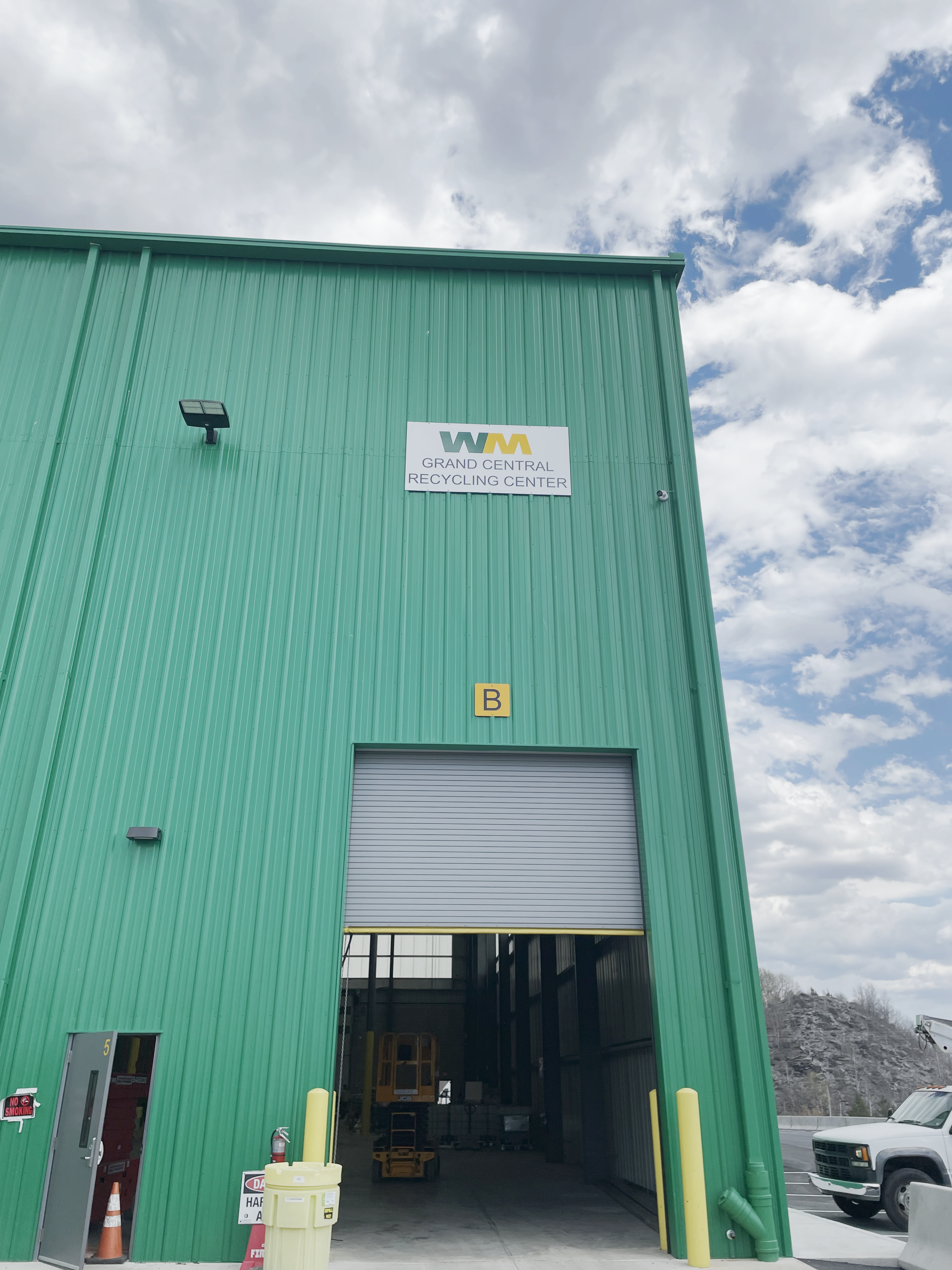 Green building with a sign that reads 'Grand Central Recycling Center' and a wide open garage door, with a parking lot and cloudy sky in the background.