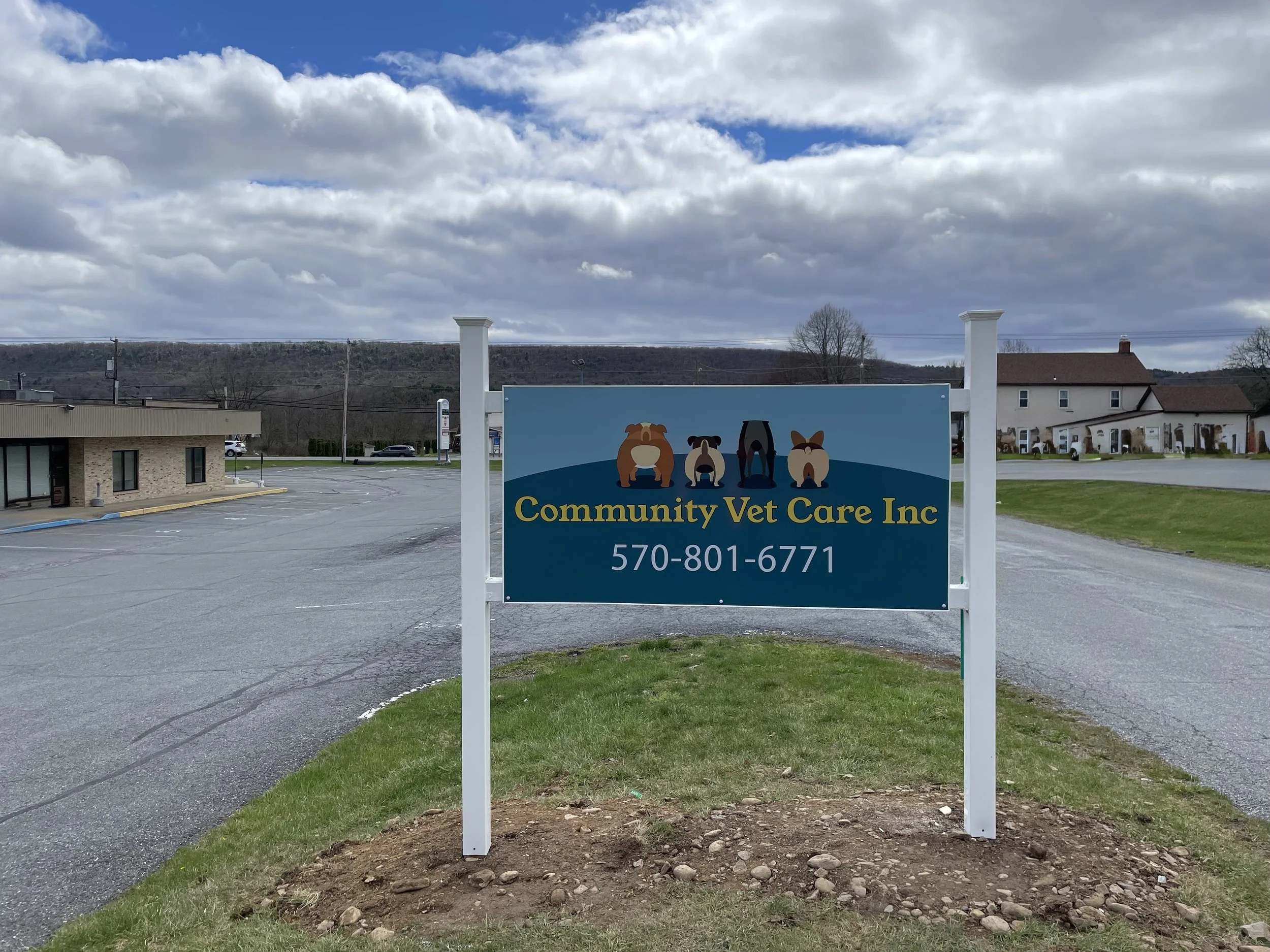 Sign for Community Vet Care Inc with illustrations of dogs and a phone number, situated on a grassy area near a parking lot.