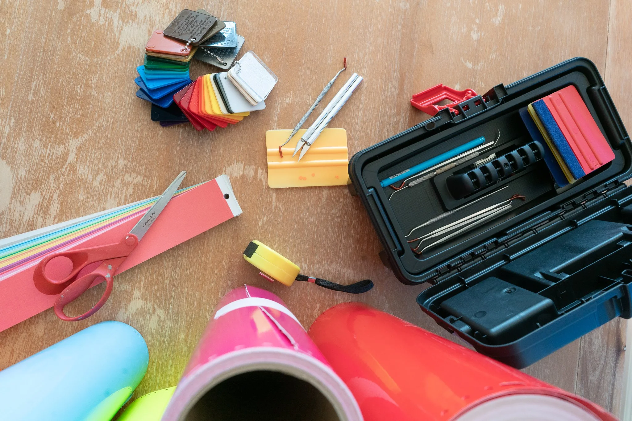A collection of craft tools and materials on a wooden surface, including scissors, color samples, a toolbox with dental tools, tape, a utility knife, and rolls of color wrapping film.