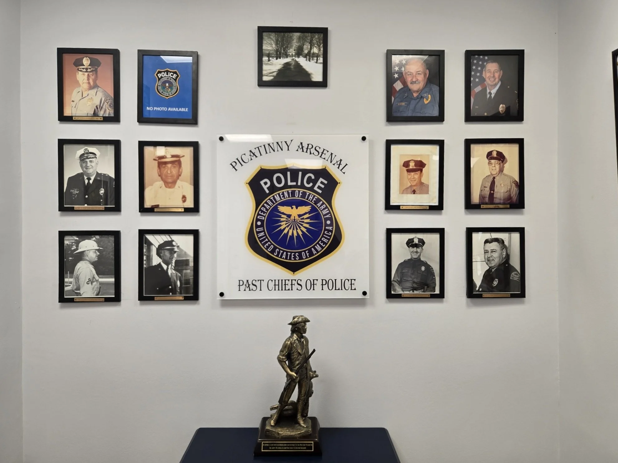 Wall display of framed police department photos and a central emblem for Picatinny Arsenal Police, past chiefs, with a bronze police statue on a table in front.