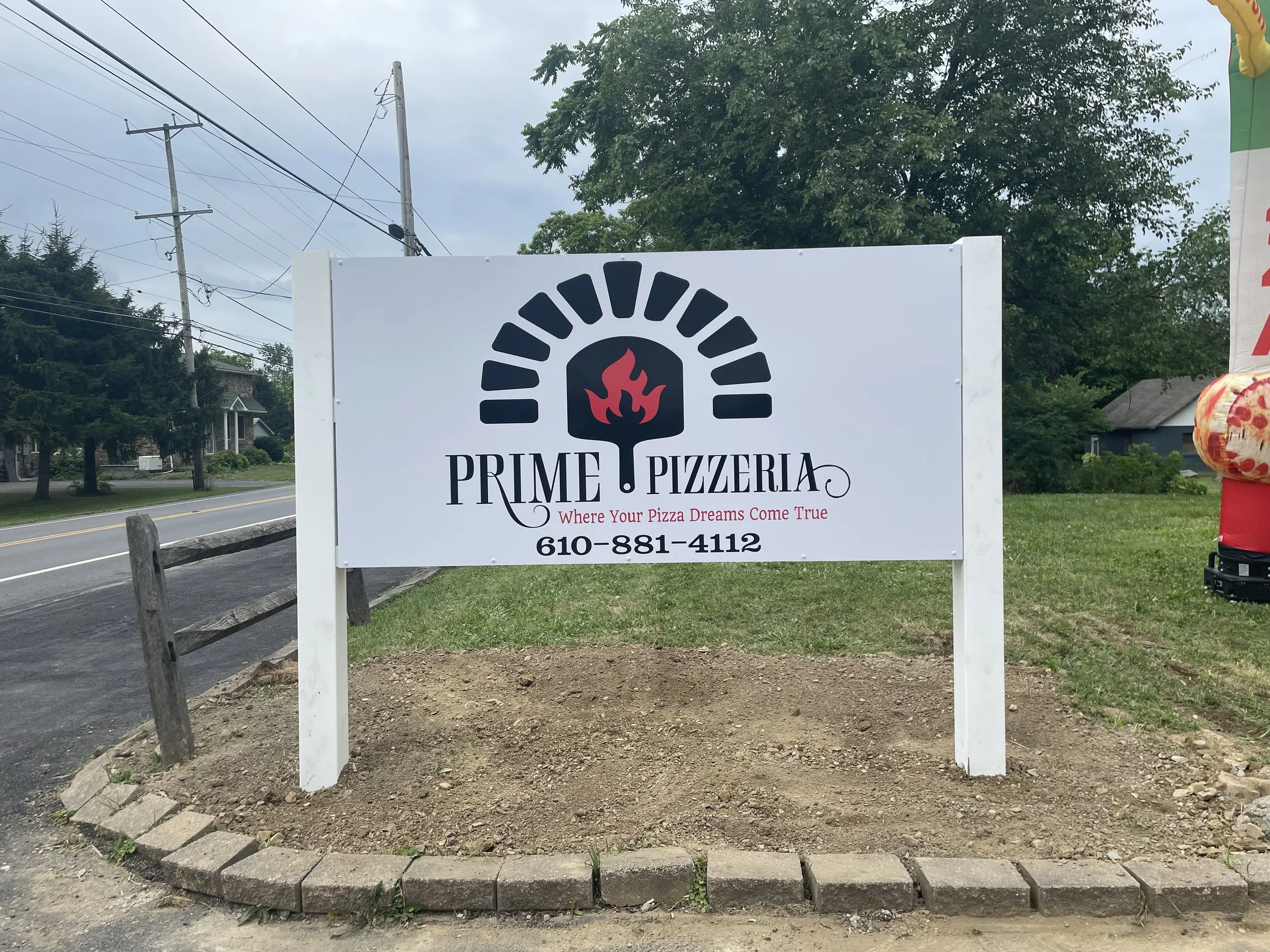 White sign for Prime Pizzeria featuring a black and red flame logo above the business name, with the slogan "Where Your Pizza Dreams Come True" and a phone number 610-881-4112.