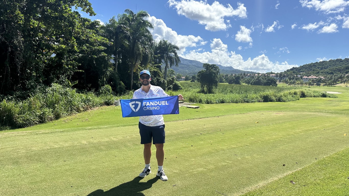 A man standing on a golf course holding a blue Fanduel Casino flag, wearing sunglasses, a hat, a floral shirt, shorts, and golf shoes with a sunny sky, trees, and mountains in the background.