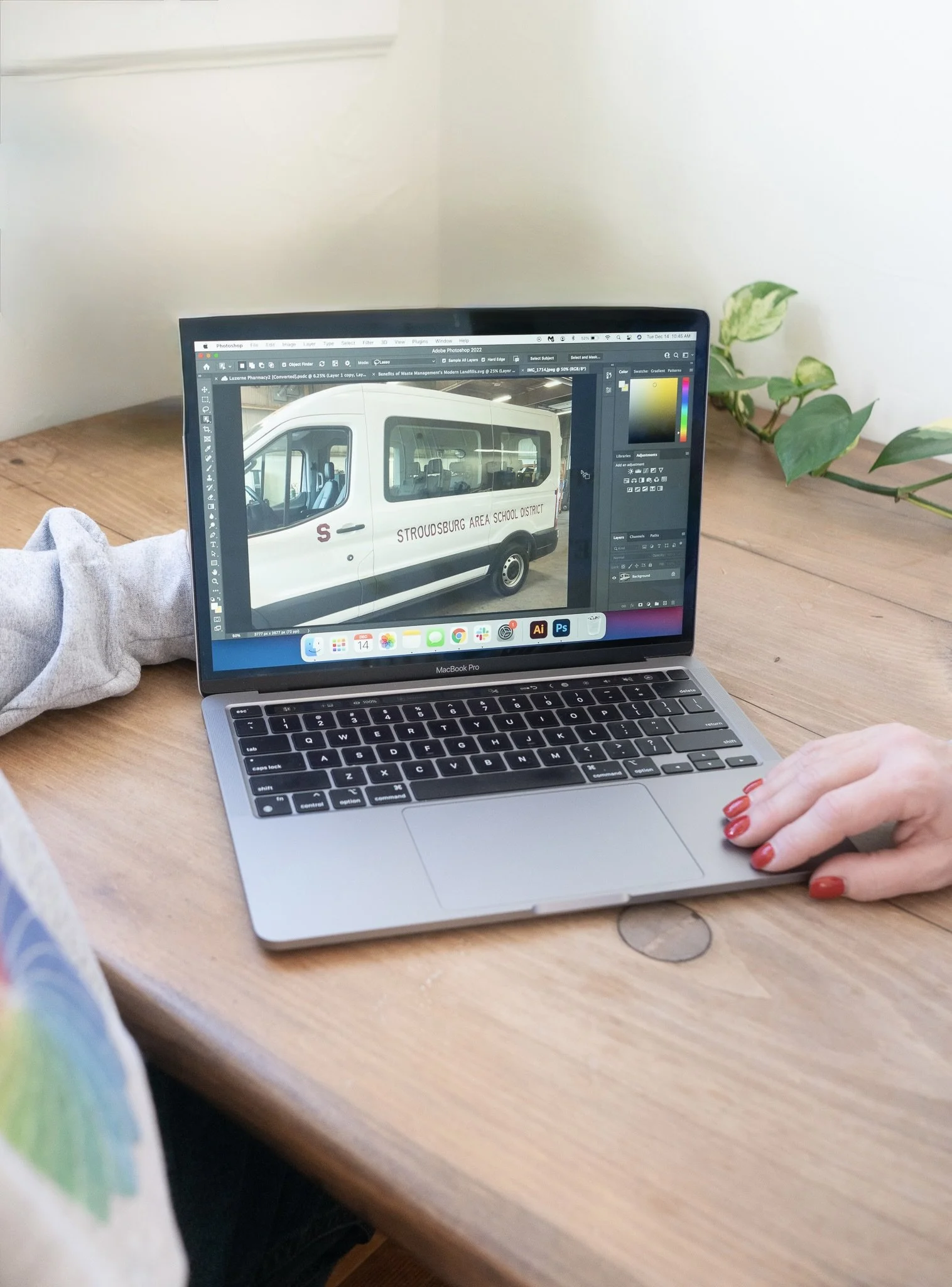 A MacBook Pro on a wooden table displaying an image of a white bus with 'Stroudsburg Area School District' written on the side. A person's hand with red nail polish is on the right side of the keyboard, and there is a gray sweatshirt sleeve on the le