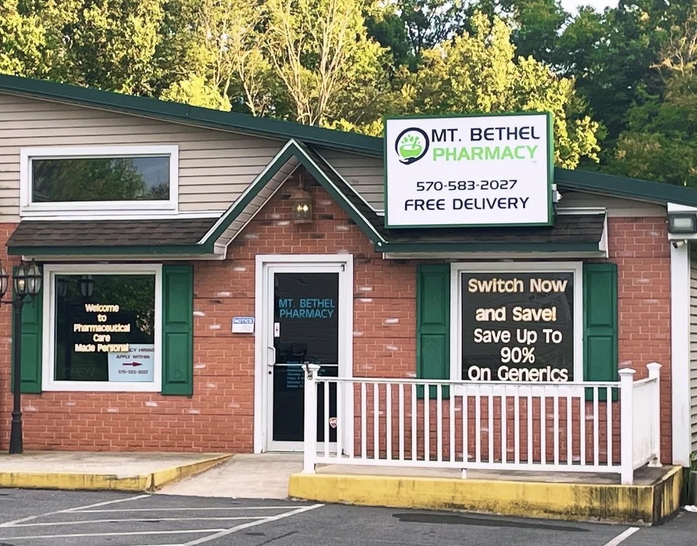 A brick pharmacy building with green shutters and a white sign that reads "MT. BETHEL PHARMACY" with the phone number 570-583-2027 and "FREE DELIVERY." There are two windows, one with a sign promoting savings on generics, and a door with a sign sayin