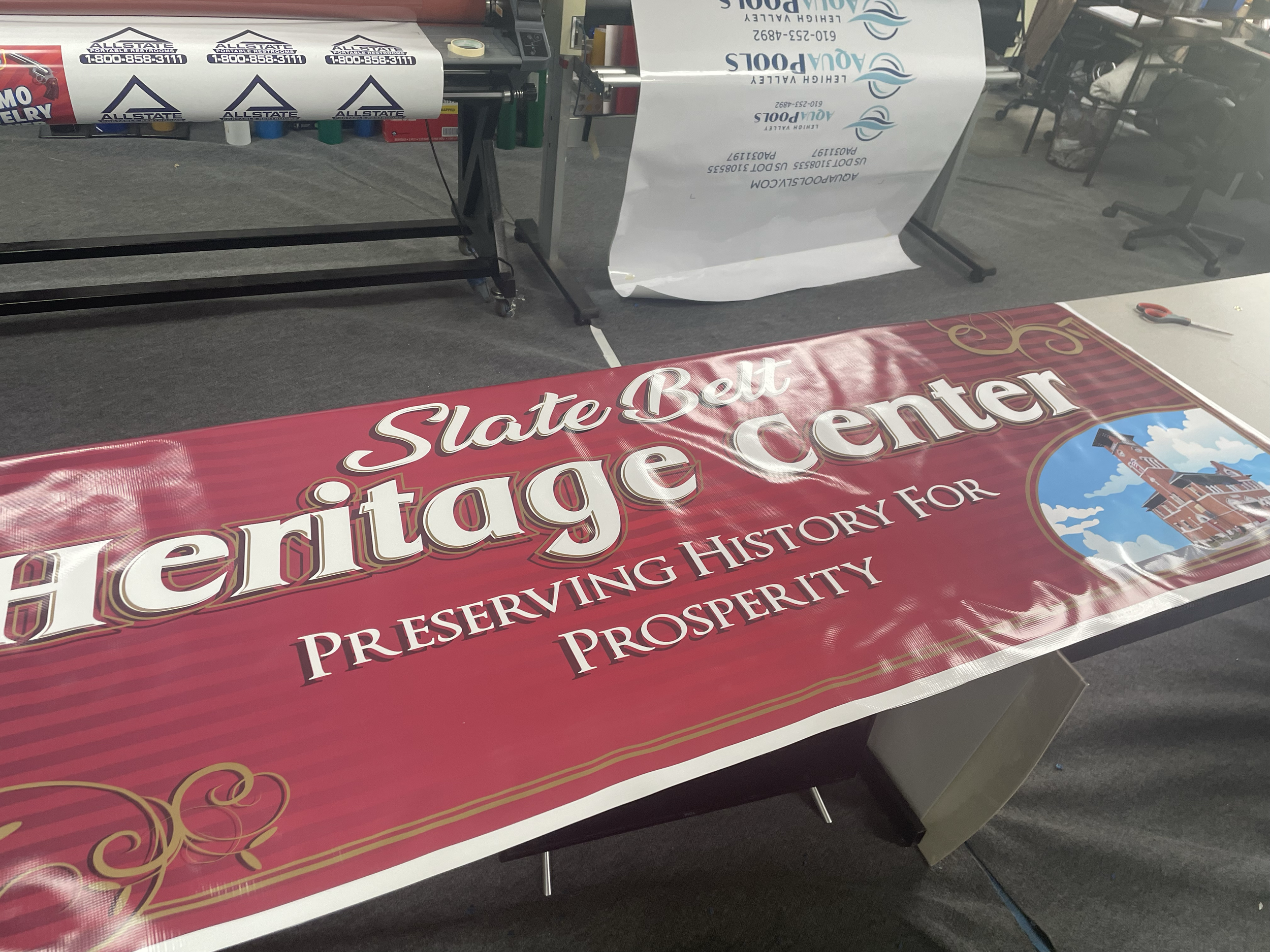 A large red banner with gold and white text that reads 'State Belt Heritage Center' and 'Preserving History for Prosperity' on a table next to scissors and a paper with printing equipment nearby.