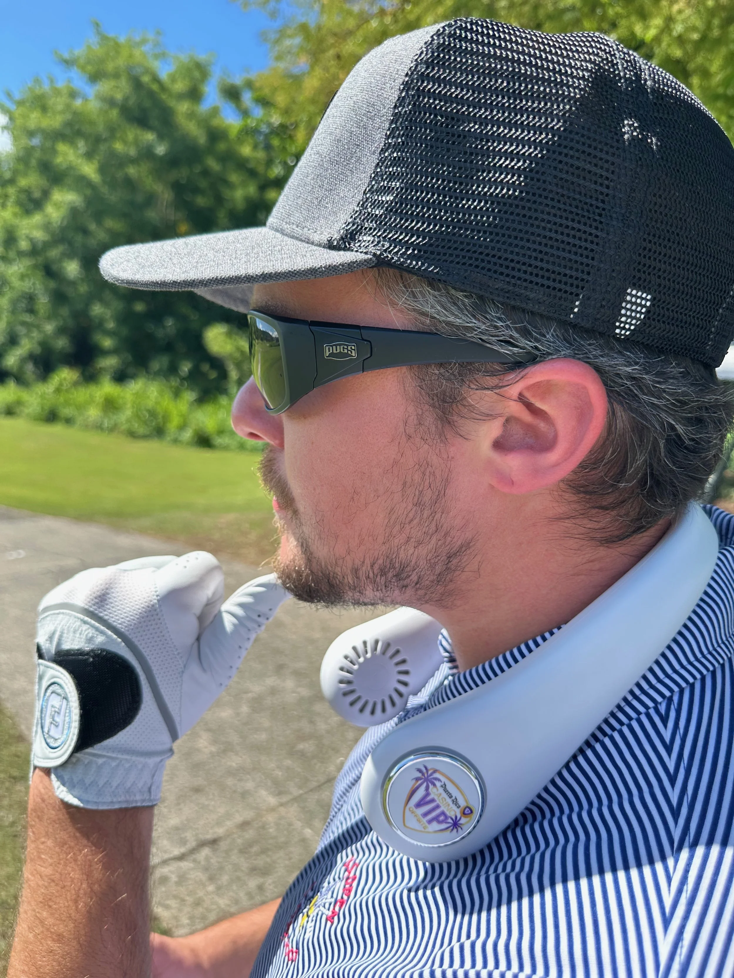 A man wearing a grey and black trucker cap, black sunglasses with the brand name PUGS, white golf gloves, and a striped blue golf shirt with a VIP logo around his neck, on a golf course with greenery and trees in the background.