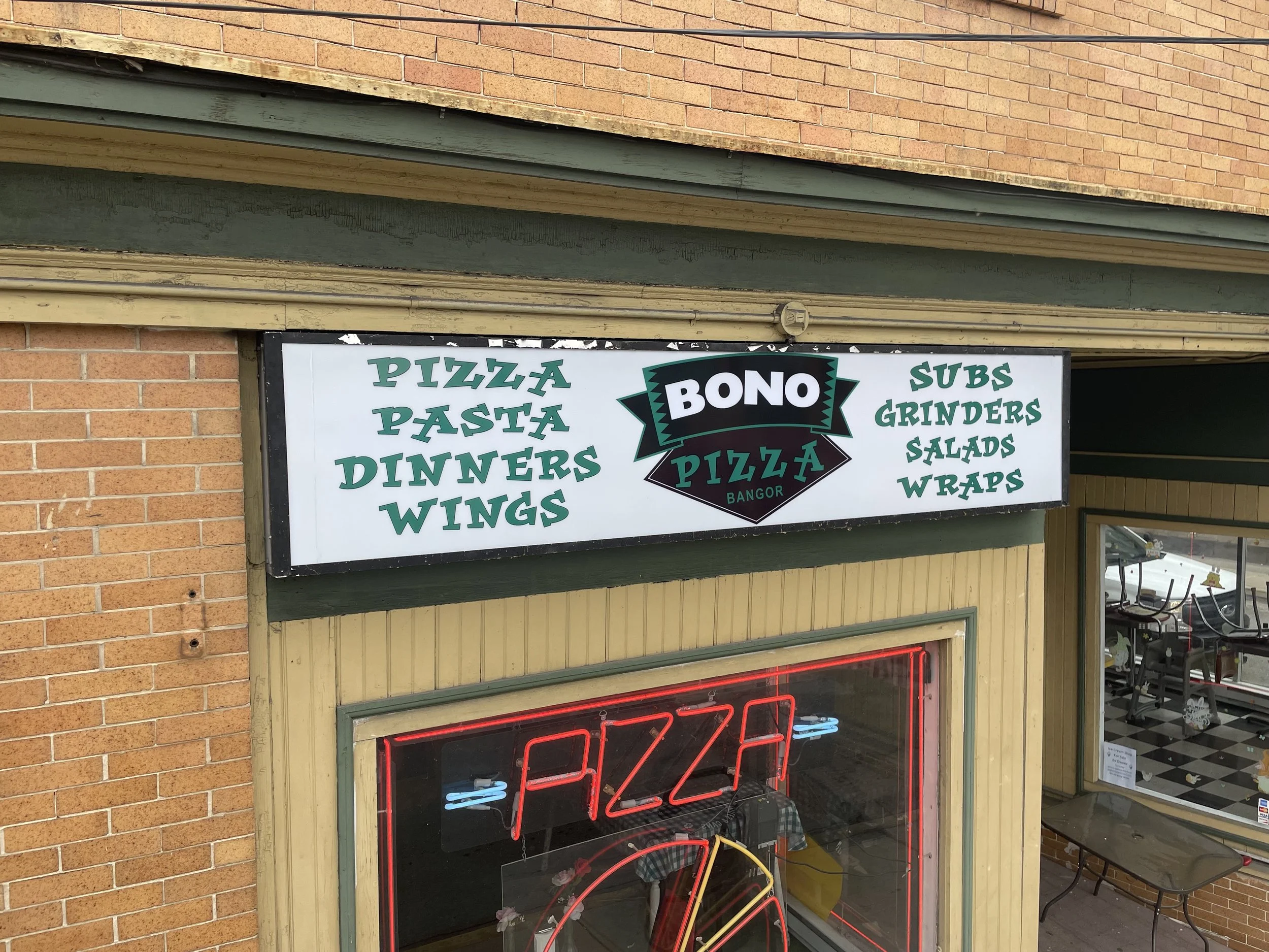 Signboard advertising a pizzeria named Bono in Bangor, listing pizza, pasta, dinners, wings, subs, grinders, salads, and wraps, with a neon sign underneath that says 'Pizza'.