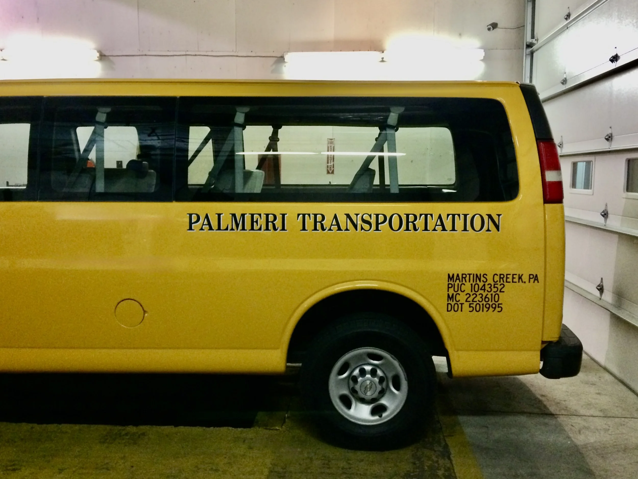 Yellow transportation van with black text reading 'PALMERI TRANSPORTATION', parked inside a garage with white walls and a closed garage door.