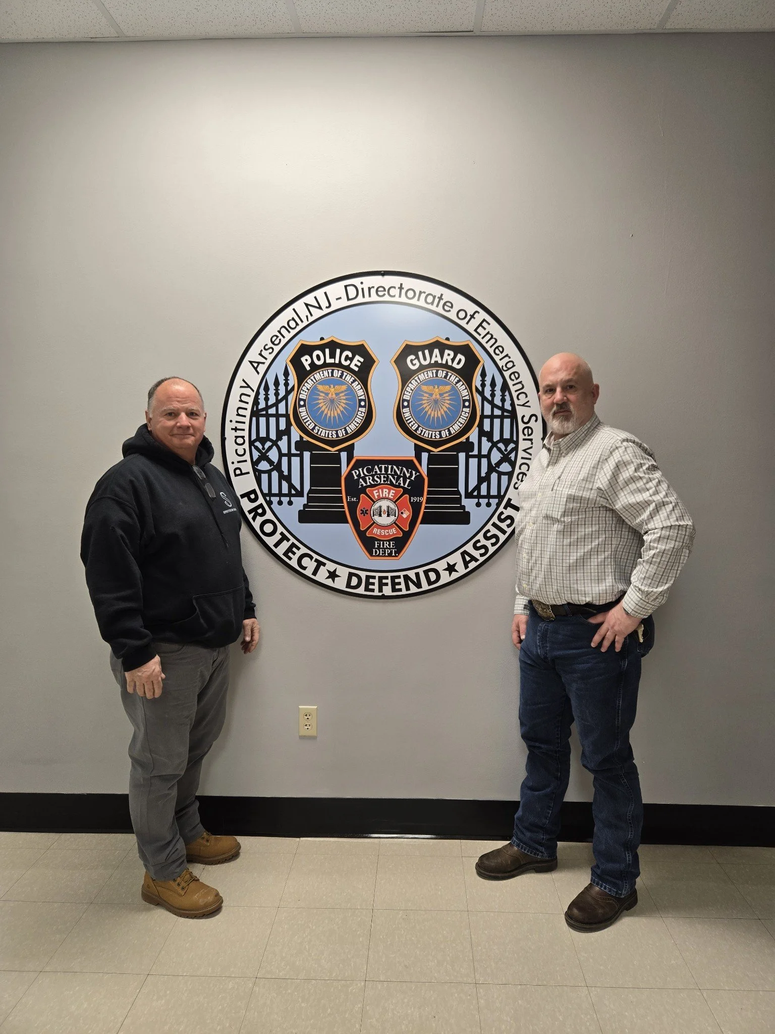 Two men standing on either side of a large emblem on a gray wall. The emblem features police shields, a fire department badge, and text indicating an emergency services alliance.