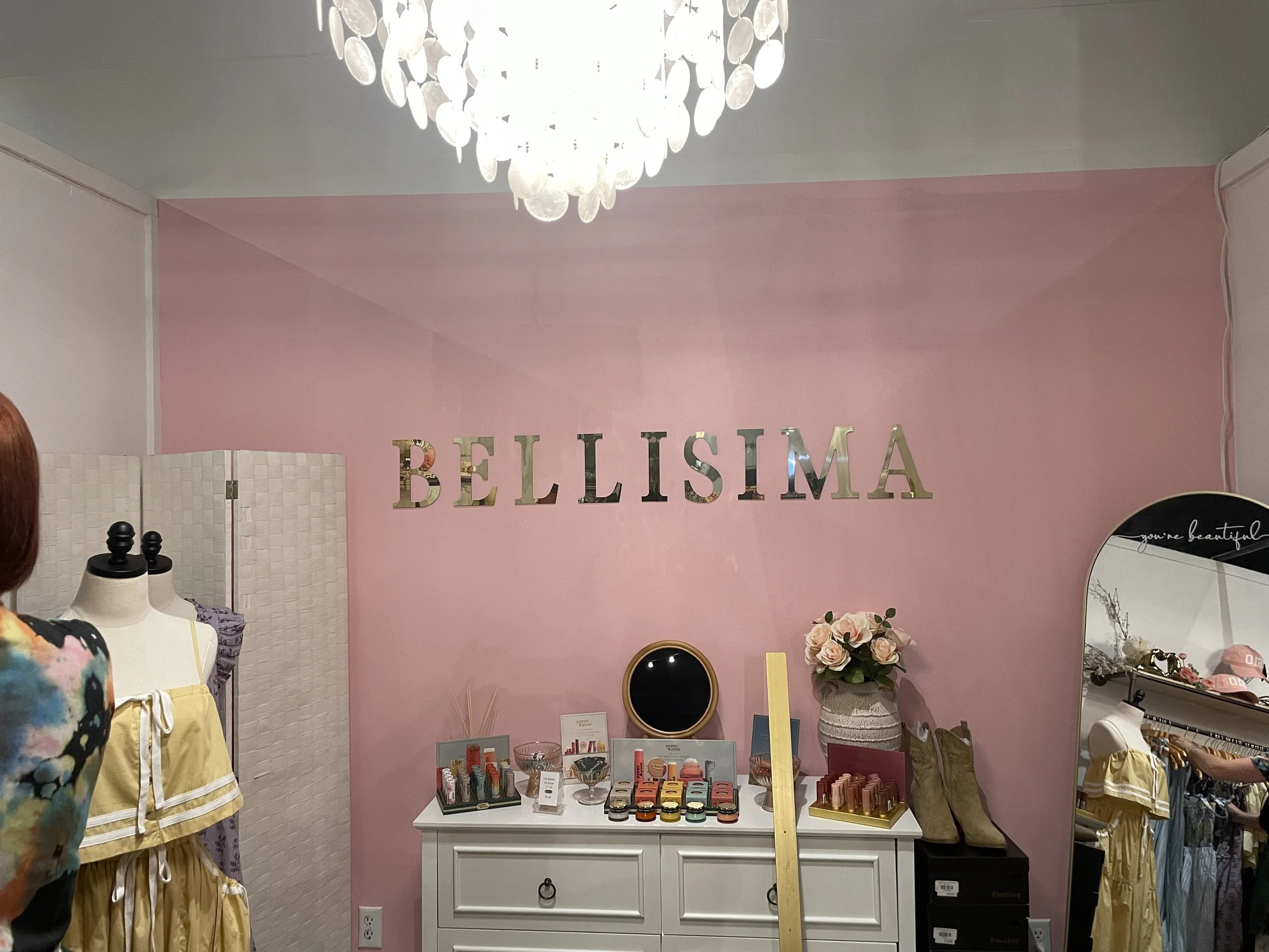 Interior of a boutique store with pink walls and a sign that reads 'BELLISIMA'. Displays of cosmetics, shoes, and accessories are arranged on a white dresser and nearby shelves, with a floral arrangement and a mirror on the dresser.