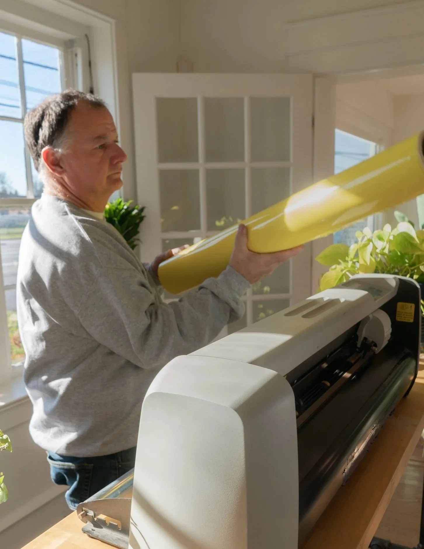 A man inspects a roll of yellow vinyl material near a settings machine, next to a window and illuminated by natural sunlight.