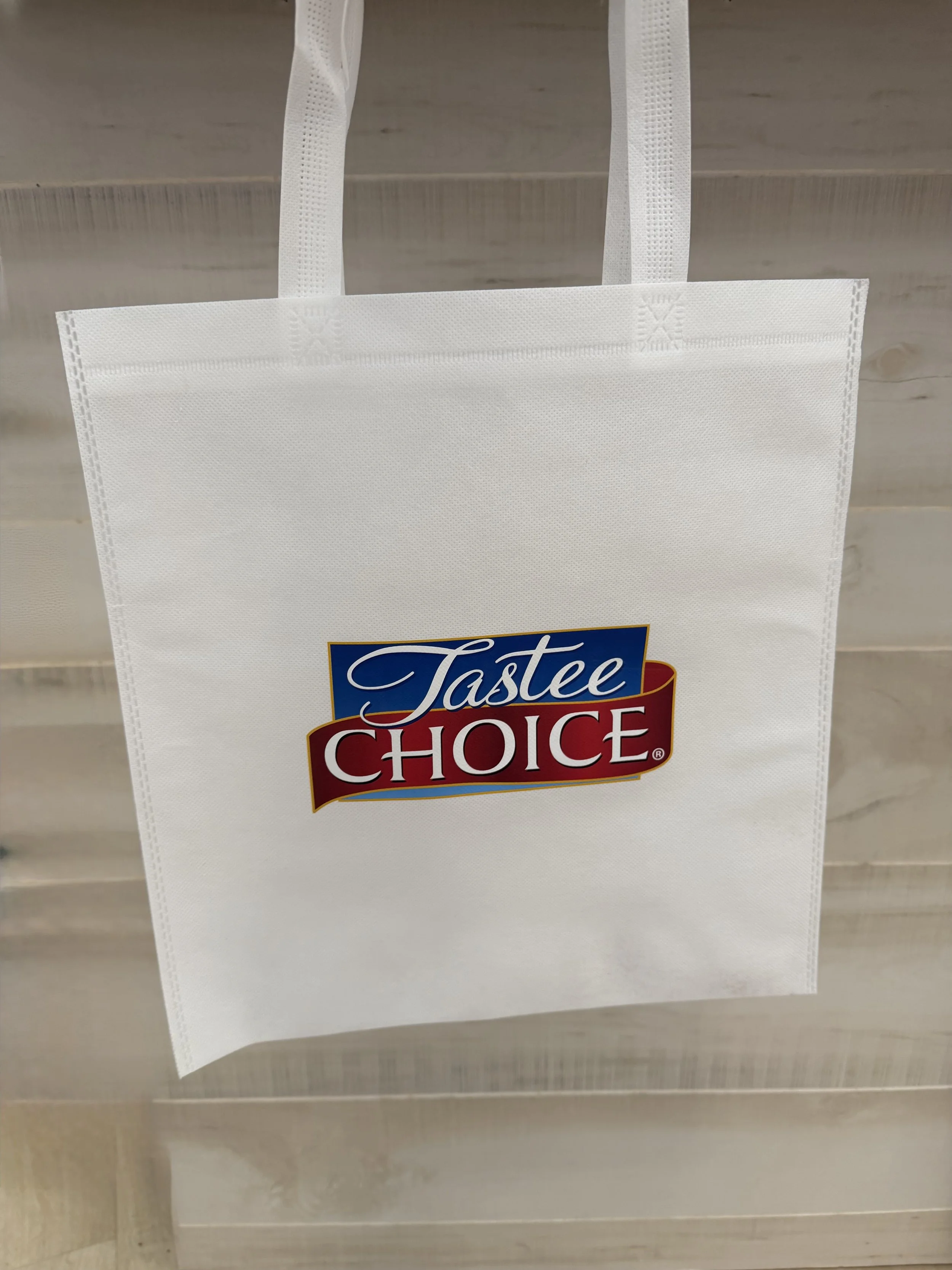 White reusable shopping bag with 'Tastee Choice' logo in blue and red, hanging against a wooden background.