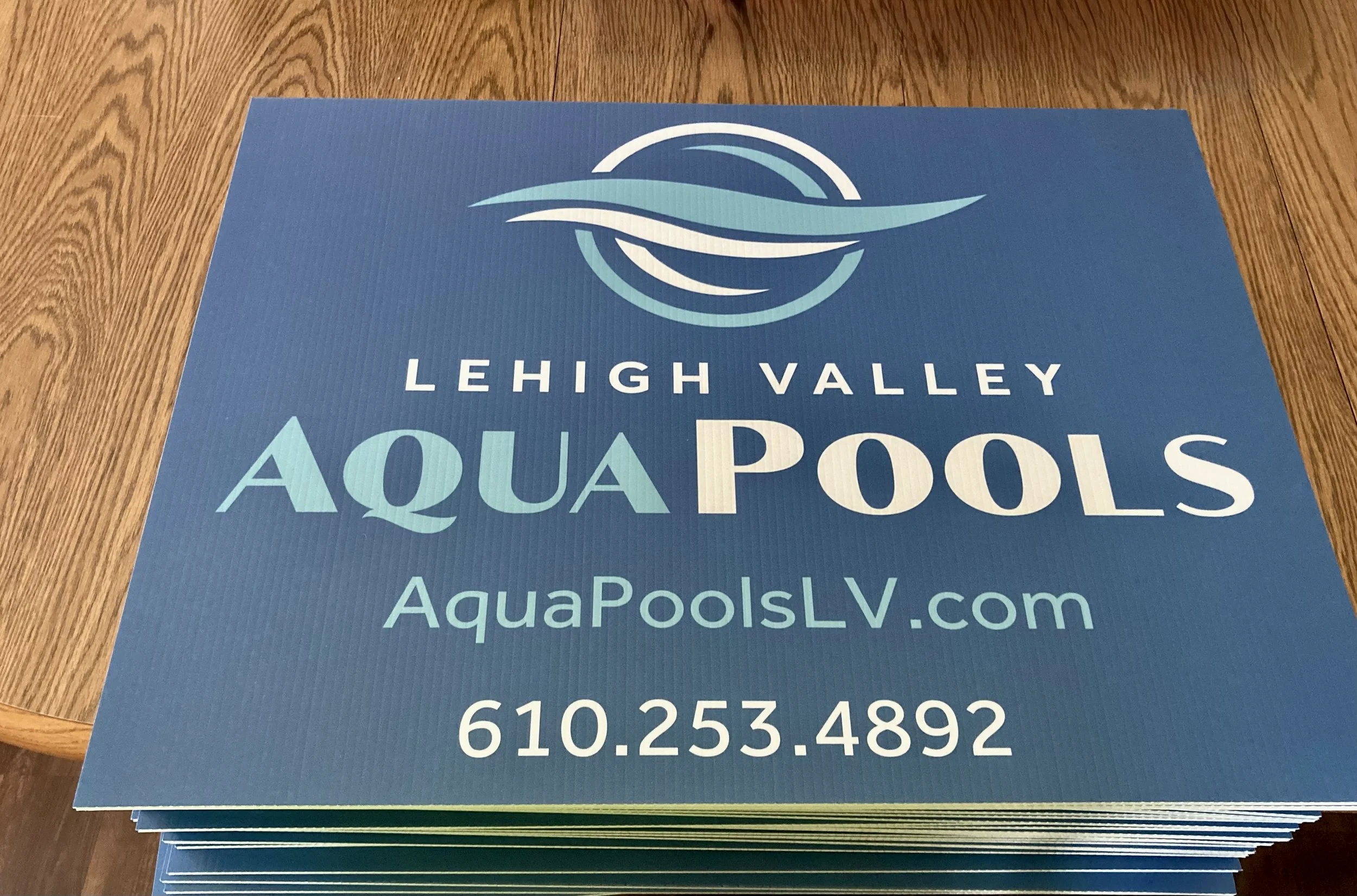 Stack of blue flyers for Lehigh Valley Aqua Pools, featuring a stylized wave logo, website AquaPoolsLV.com, and phone number 610-253-4892, on a wooden surface.
