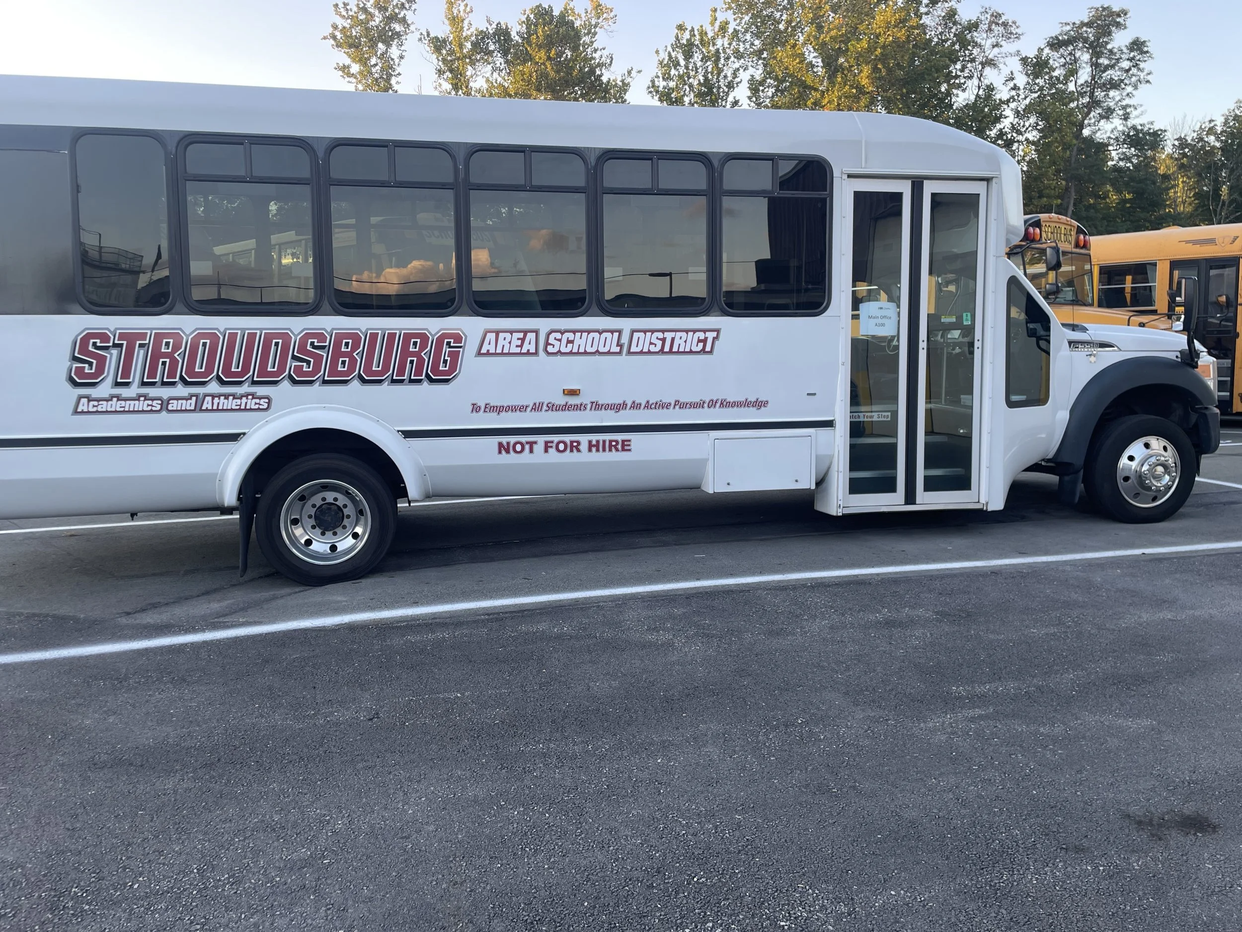White bus parked in a lot with school buses in the background. The bus has the words 'Stroudsburg Area School District' and 'Academics and Athletics' printed on the side, along with a slogan and a note that it is 'Not for Hire.'