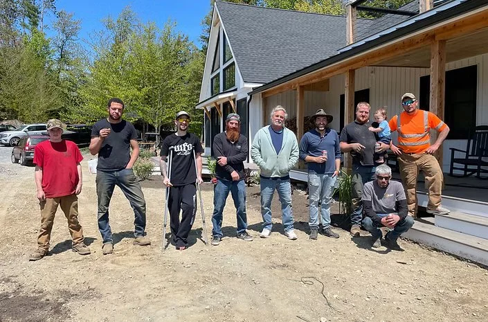 Group of nine people standing outside a house with a porch, some holding drinks, with trees and parked cars in the background.