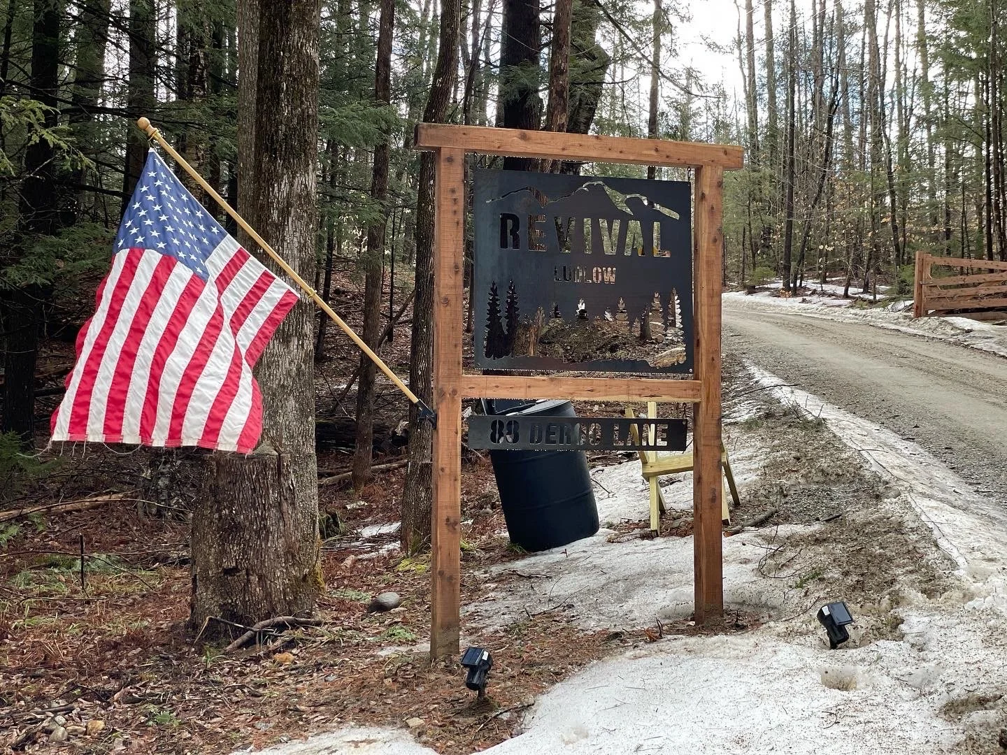 A sign reads 'Revival Hollow' and '08 Demo Lane' in a wooded area with trees, a dirt road, and a partially snow-covered ground. An American flag is attached to a tree nearby.