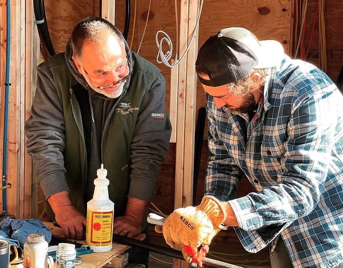 Two men working together in a woodworking shop, one wearing glasses and a dark vest, the other wearing a plaid shirt, a backwards cap, and gloves, examining a piece of wood.