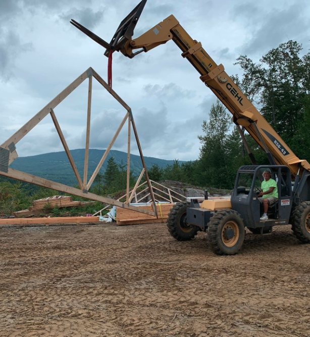 A construction worker operating a large yellow crane lifts a wooden truss into place for building. The construction site is outdoors with trees, mountains, and a cloudy sky in the background.