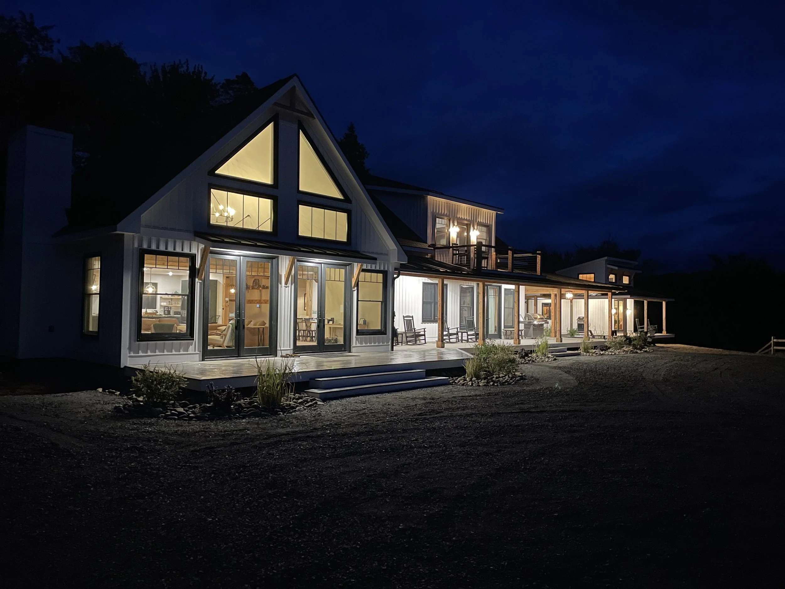 A modern house at night with interior lights on, featuring large windows and a covered porch with chairs.