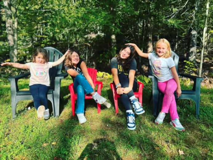 Four young girls sitting outdoors on chairs, smiling and playing in a wooded area.