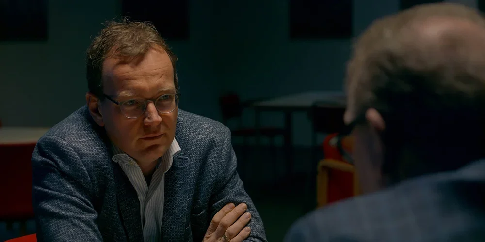 A man wearing glasses and a blue blazer sitting at a table and talking to another man whose back is to the camera.