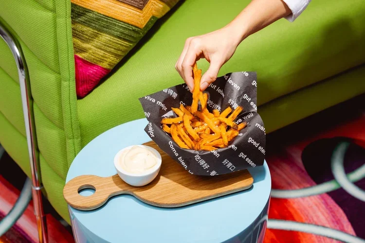 A hand reaching into a black paper-lined basket of sweet potato fries on a small round table, with a small bowl of dipping sauce on a wooden board next to it.