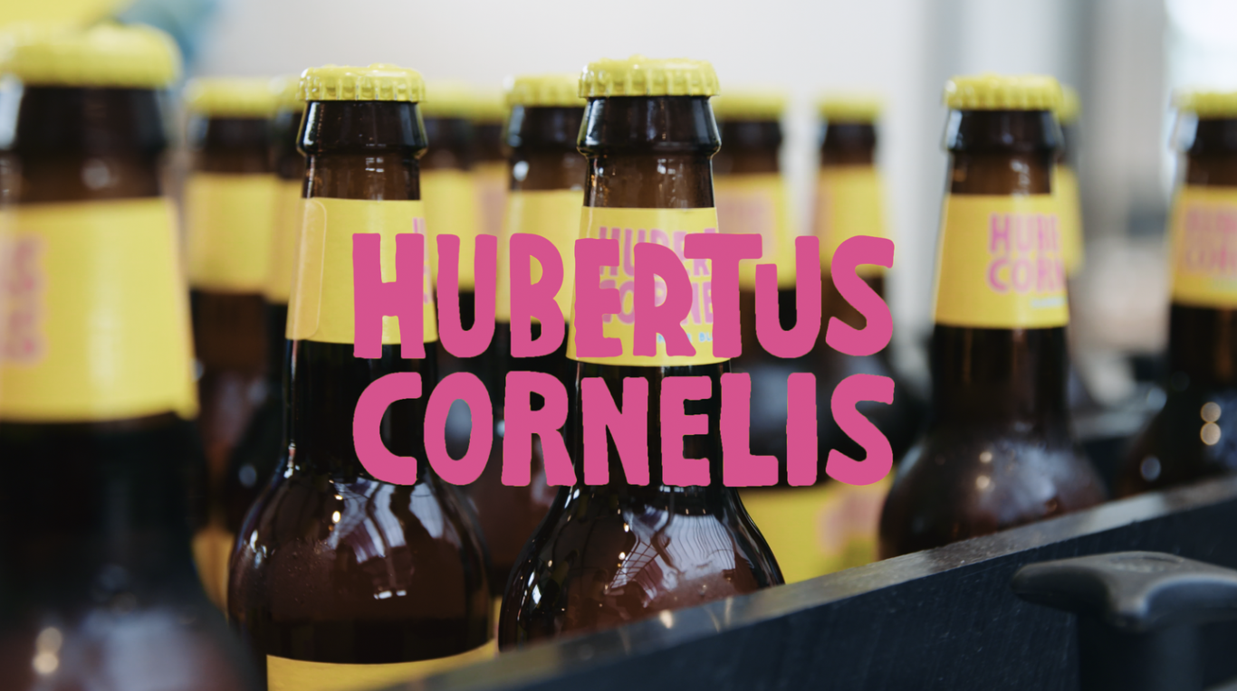 Close-up of bottles with yellow labels and yellow caps, with the text 'HUBERTUS CORNELIS' in large pink letters overlaying the bottles.