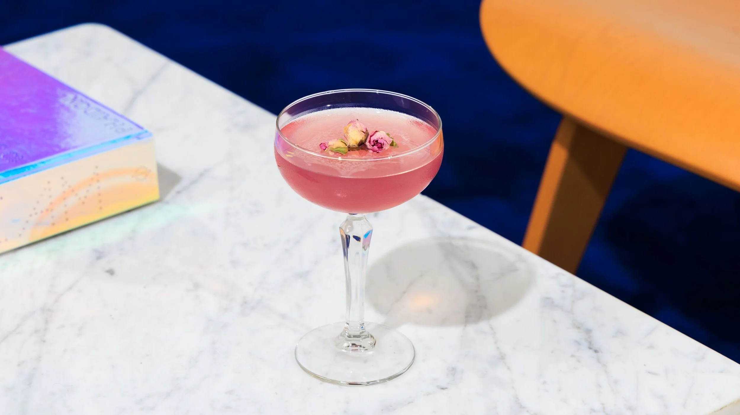 A pink cocktail in a crystal stemmed glass garnished with small pink flowers on top, placed on a white marble surface.