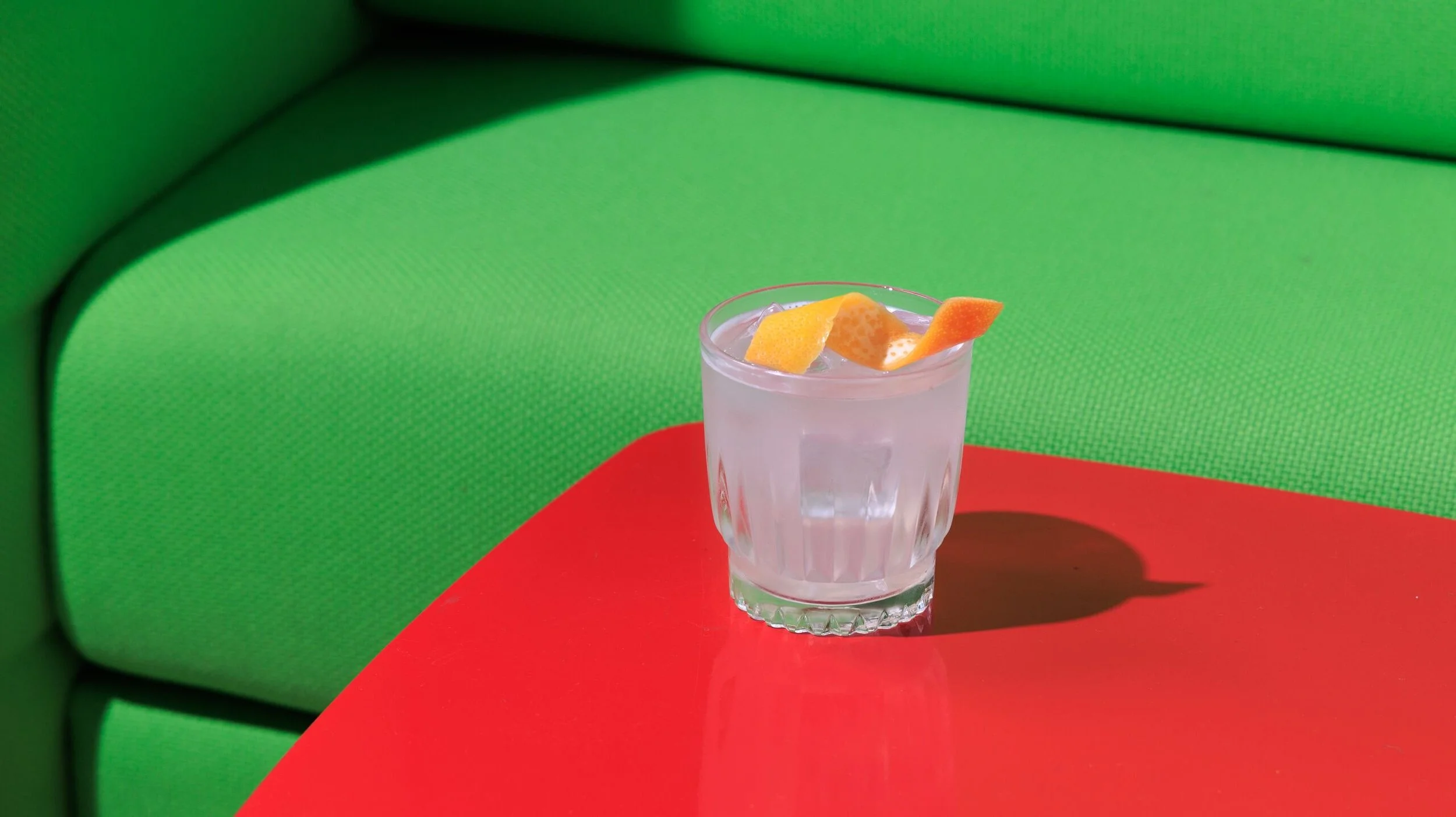 A glass of clear liquid with orange peel garnish on a red table against a green background.