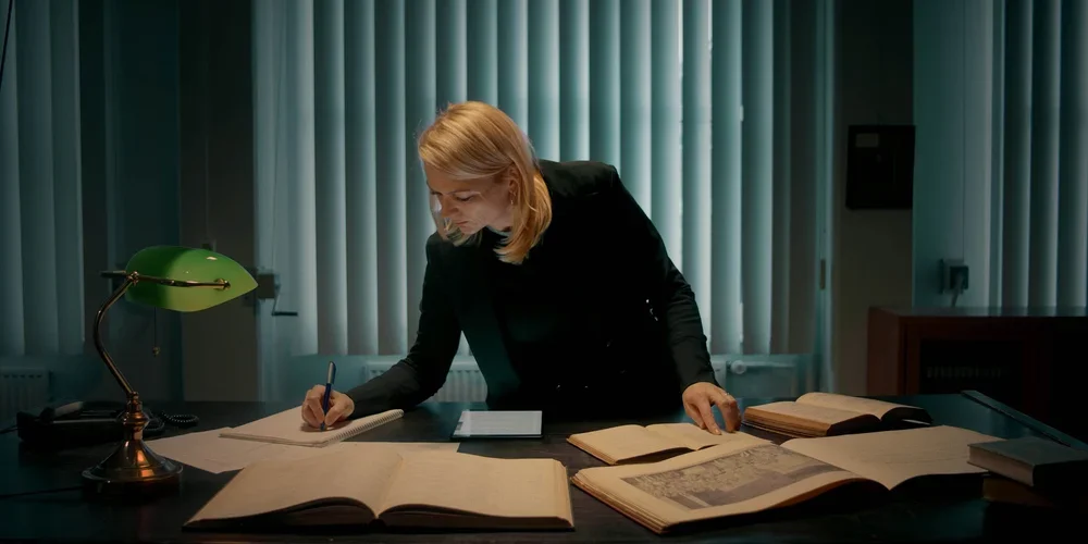 Woman with blonde hair working at a desk filled with large open books and notebooks, lit by a green desk lamp, in a room with green curtains.