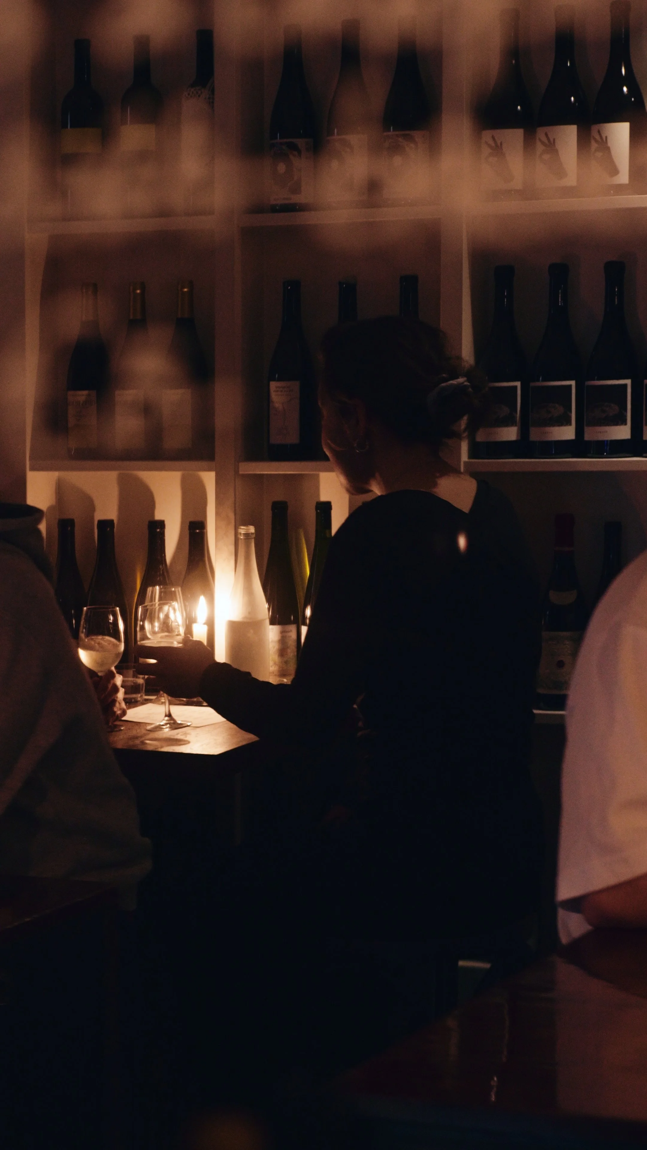 A woman in a dark dress holding a glass of wine, standing at a table in a dimly lit wine cellar or bar with shelves of wine bottles behind her.