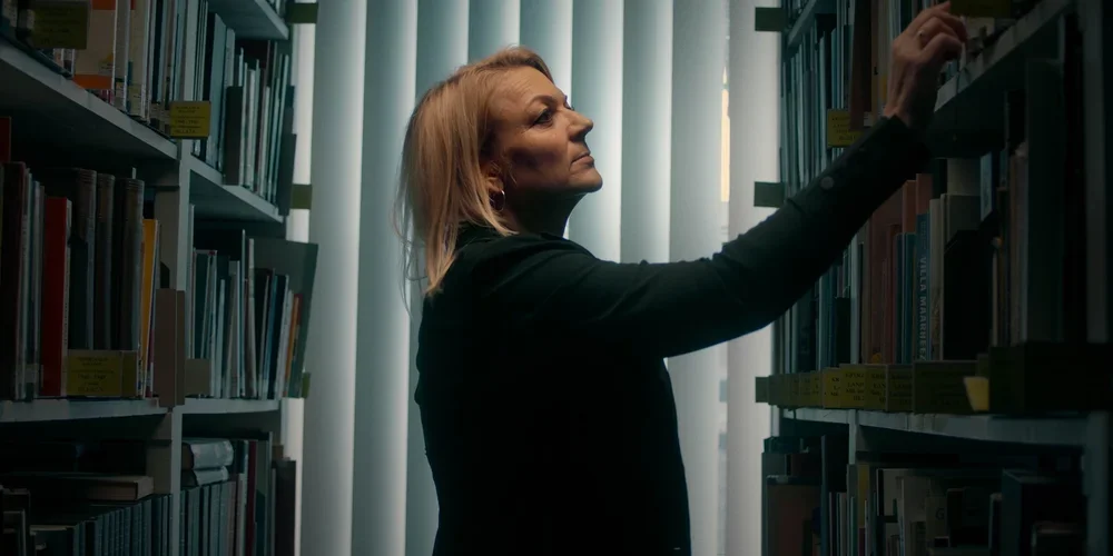 A woman with blonde hair in a library, reaching for a book on a shelf, with light coming from behind through curtains.