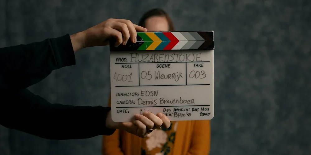 A person holding a slate with production details in front of a woman wearing an orange jacket.