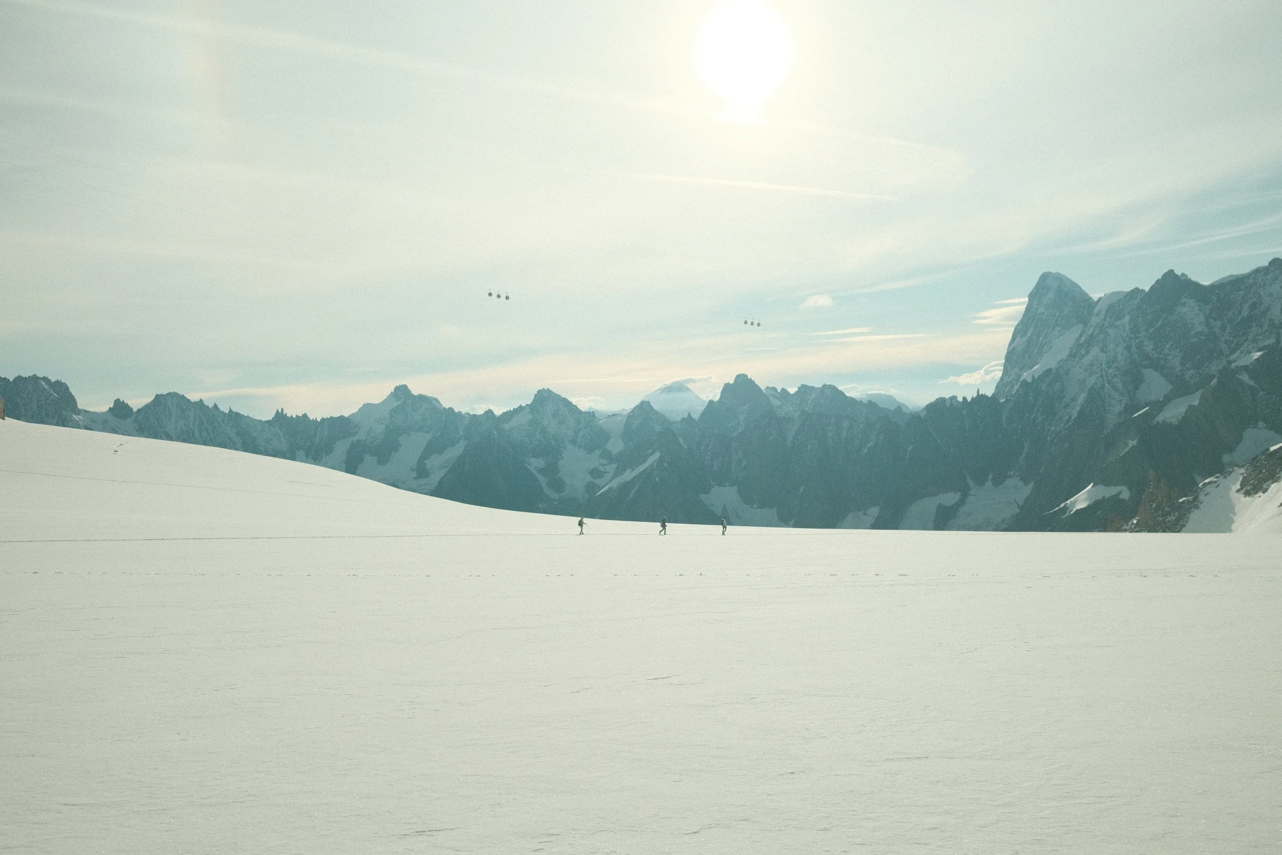 Snow-covered mountain landscape with three skiers walking across a vast snowfield, mountain peaks in the background, and a partly cloudy sky.