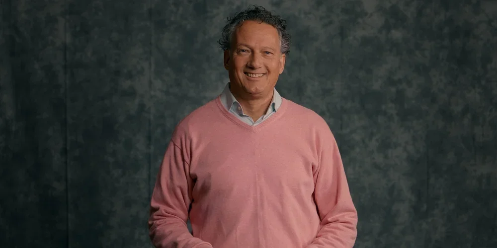A smiling middle-aged man with curly hair wearing a pink sweater and a collared shirt standing in front of a dark textured background.