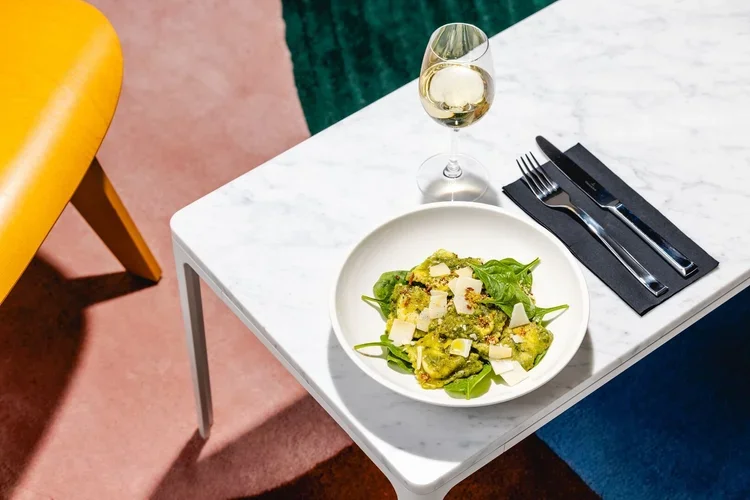 A white marble table with a bowl of green salad topped with cheese, a glass of white wine, and black cutlery on a black napkin.