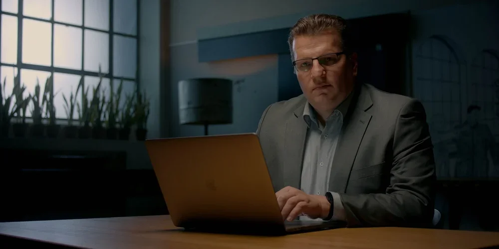 A man in a gray suit and glasses working on a laptop in an office with a window and plants in the background.