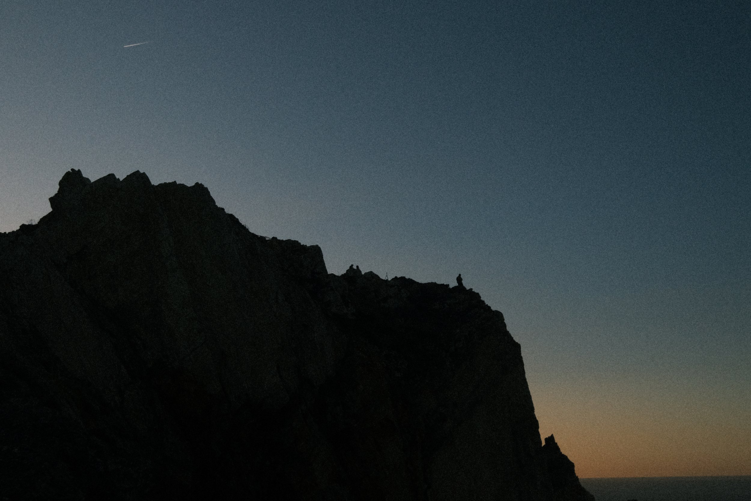 Silhouette of a person standing on a mountain ridge at dusk or dawn, with a fading light sky and faint contrail visible.