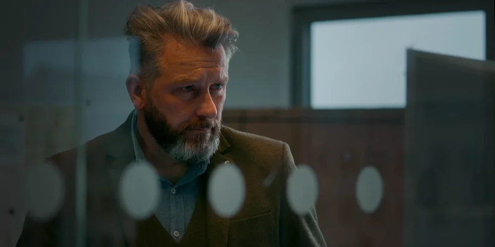 A serious middle-aged man with a beard and gray hair, dressed in a blazer and collared shirt, sitting in front of a computer screen in a modern office or conference room, with a window in the background.