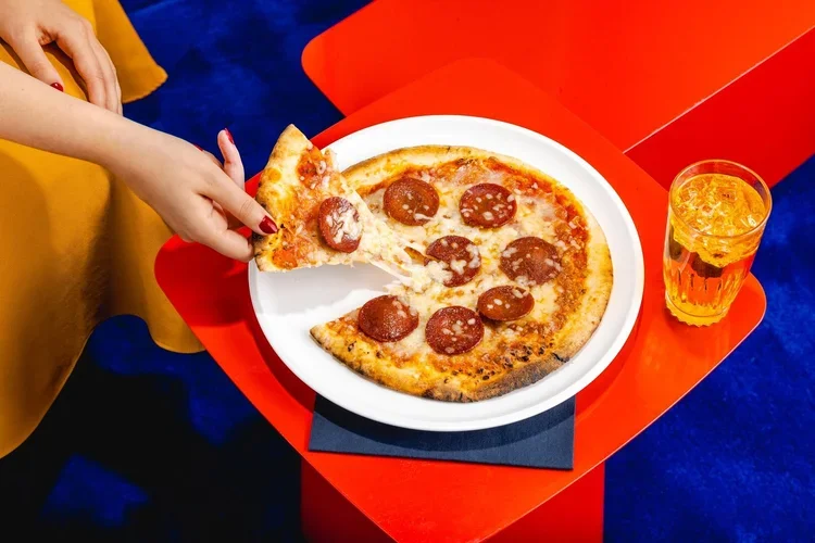 People serving themselves pepperoni pizza from a white plate and a glass of orange drink on a red table.