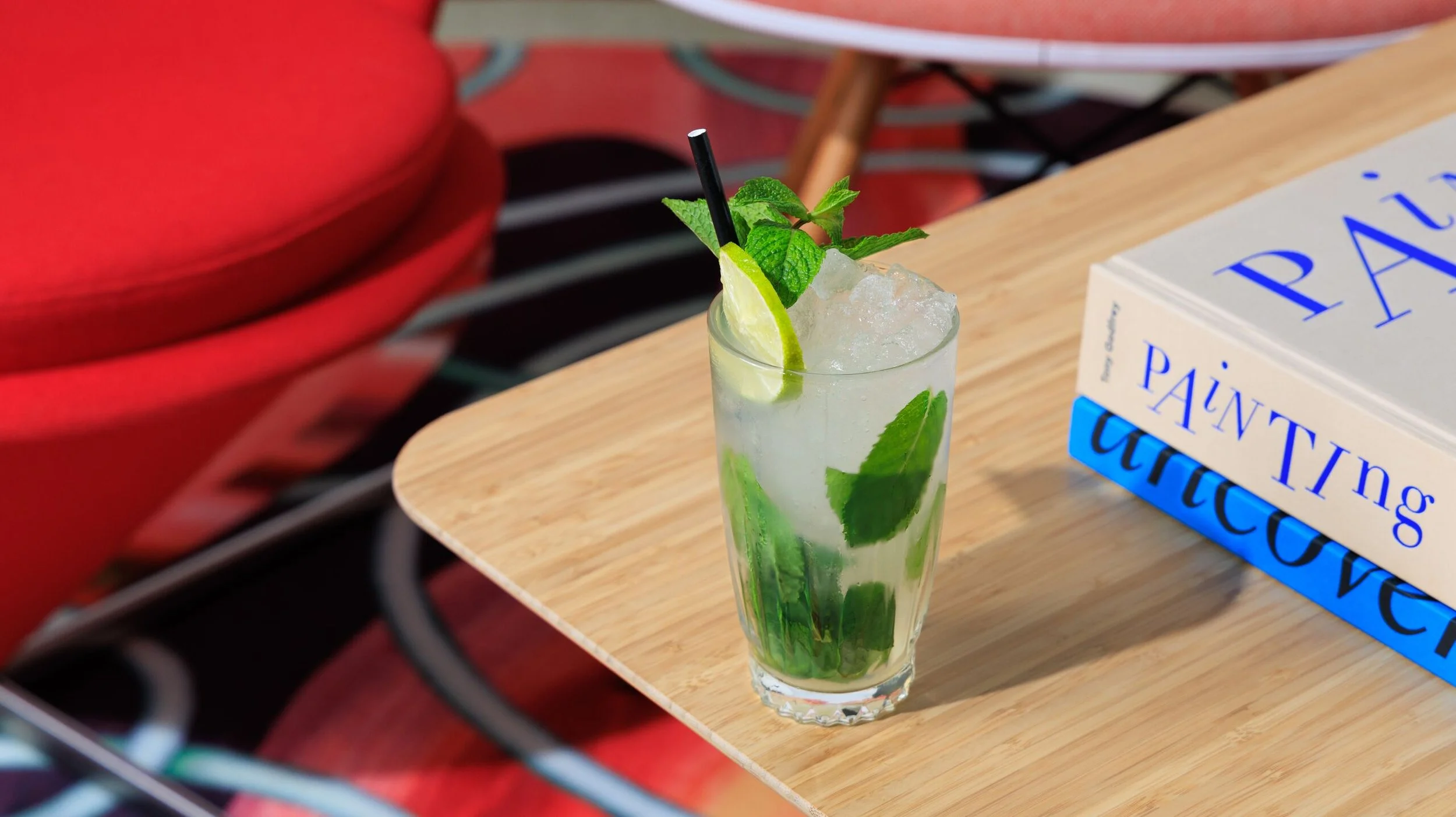 A glass of mojito with lime wedge, mint leaves, and ice on a wooden table next to a book titled 'Painting the Universe'.