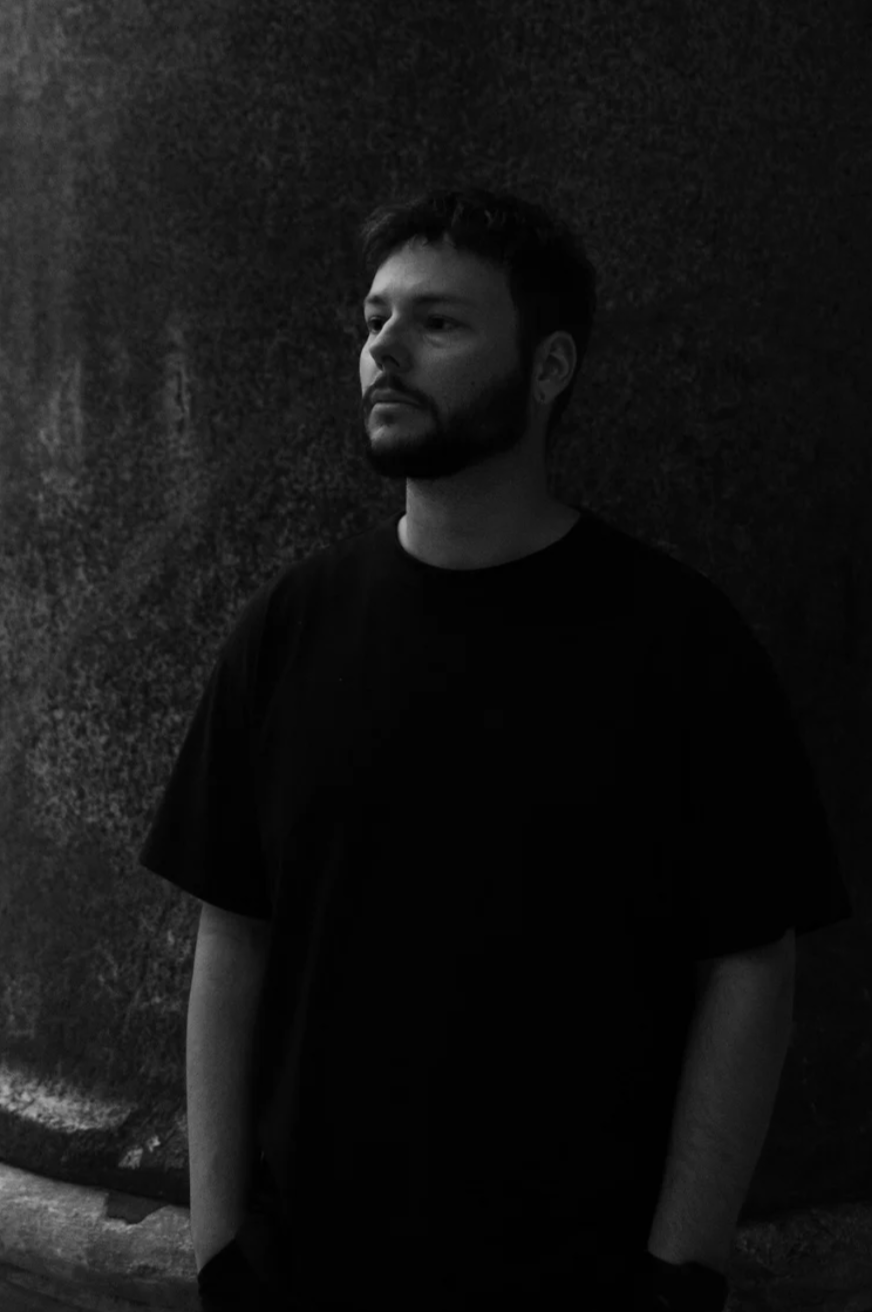 A young man with a beard and mustache in a black t-shirt stands against a textured dark wall, looking to the side in black and white.