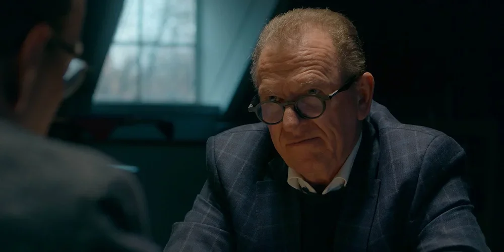 A middle-aged man with glasses and gray hair sits at a table, talking to someone whose back is to the camera, in a dimly lit room with a window in the background.