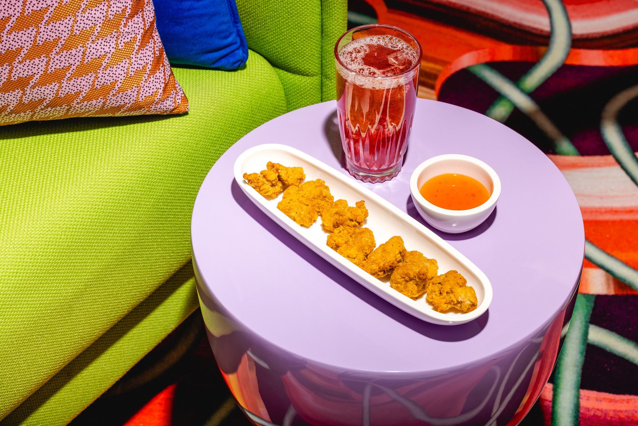 A purple table with a white platter of fried chicken pieces, a small bowl of dipping sauce, a glass of pink drink, and a small container of orange sauce, next to a green couch with colorful pillows and a multicolored carpet.