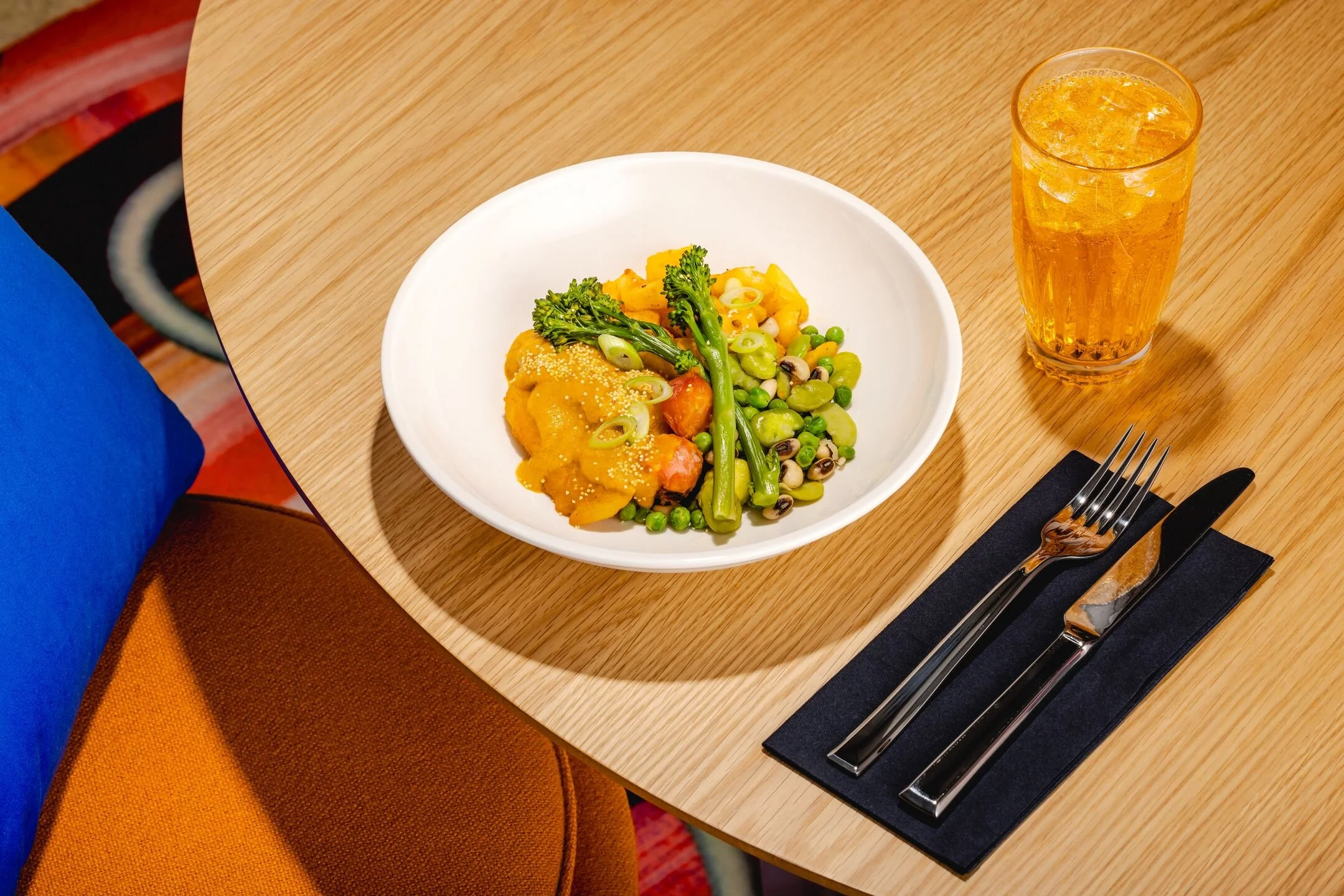 A plate of vegetable casserole with broccoli, peas, carrots, and squash, served on a white dish. A glass of iced tea or soda is next to it. The setting includes a fork and knife on a black napkin on a wooden table.