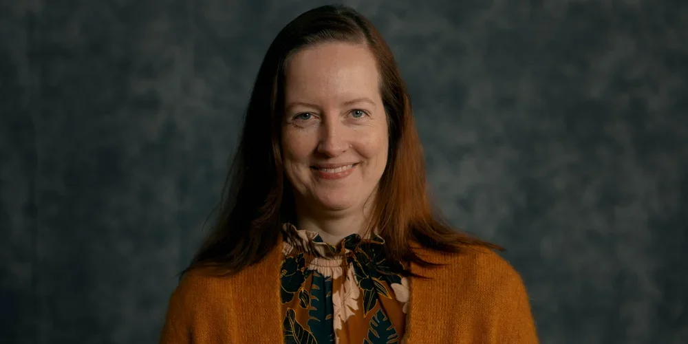 A woman with long red hair smiling in front of a dark, blurred background.
