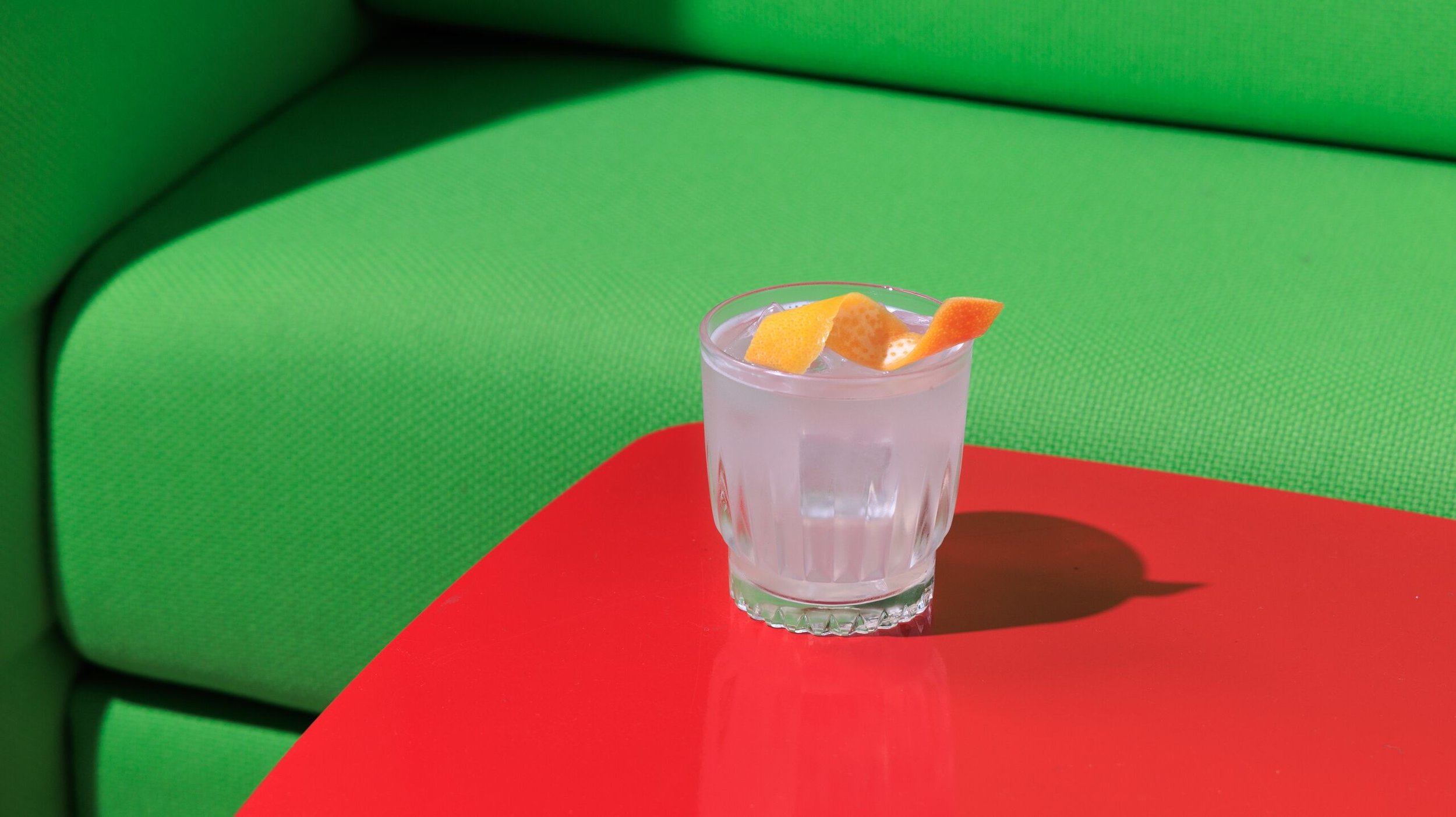 A clear glass of lemonade with ice and lemon peel garnish on a red table, with a green textured background.